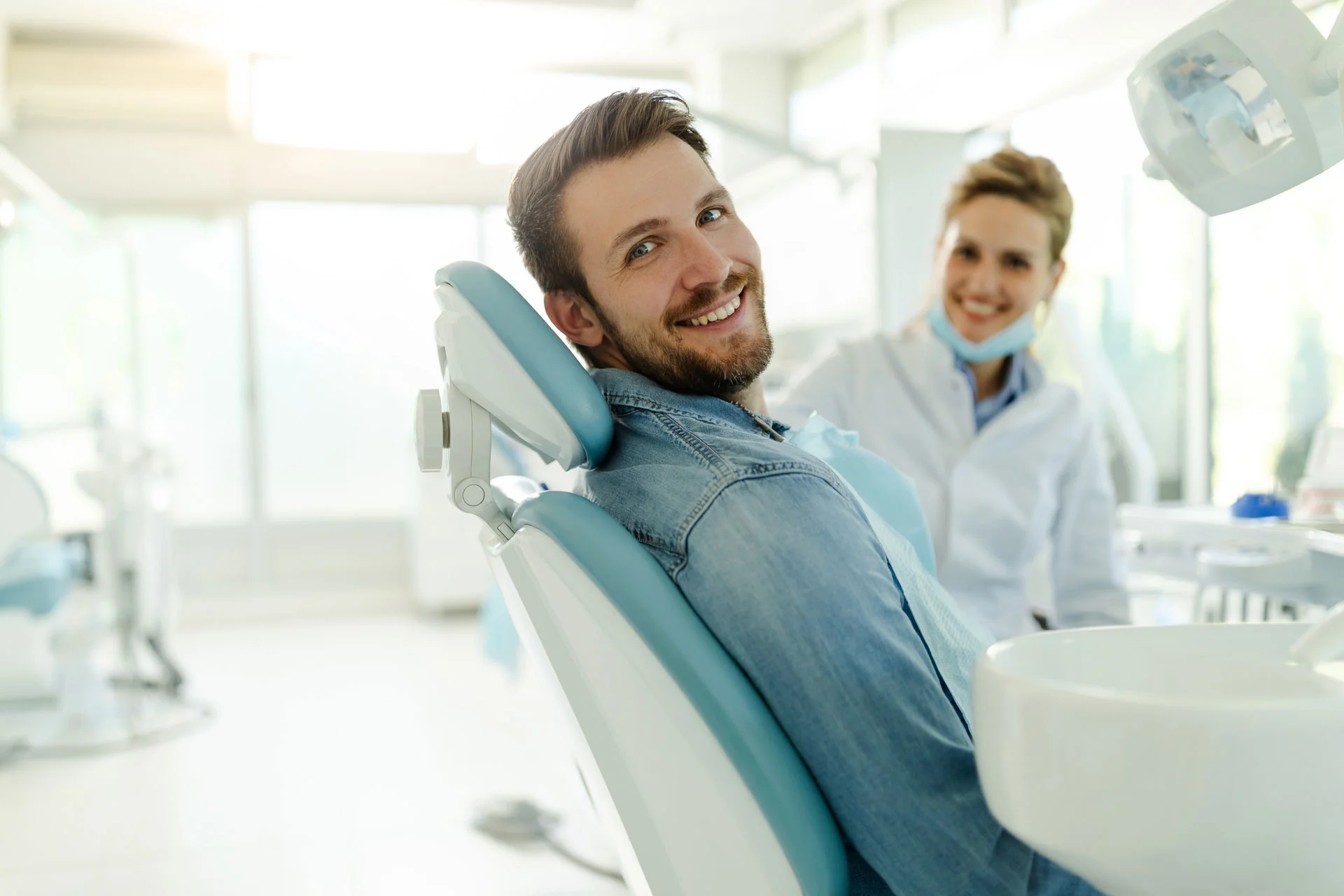 Smiling male patient sitting in a dental chair while a dentist prepares equipment in a bright, modern clinic.